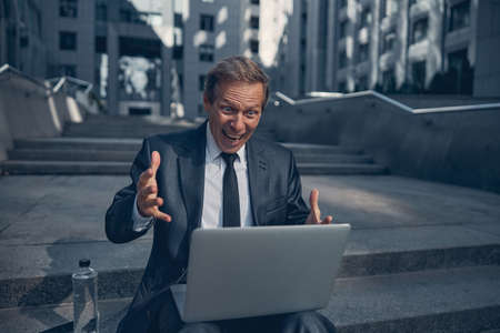 Cheerful excited man in elegant suit looking at notebook display with amused expression while...