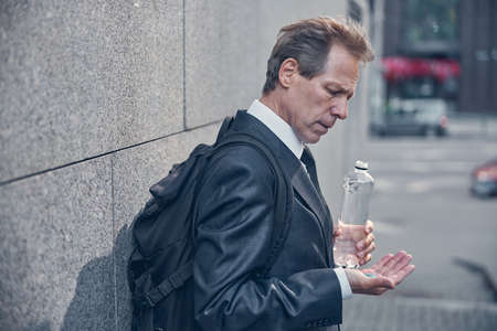 Handsome gentleman with backpack looking at blue capsule on his hand while standing on the street