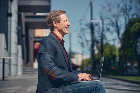Handsome stylish gentleman looking away and smiling while using modern notebook outdoorsの写真素材