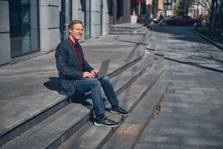 Handsome gentleman looking away and smiling while sitting on steps and working on notebookの写真素材