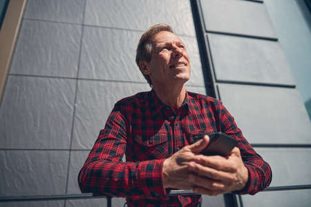 Cheerful gentleman in shirt holding smartphone and smiling while spending time outdoors