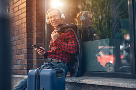 Handsome gentleman in headphones holding mobile phone and smiling while enjoying favorite songs on the streetの写真素材