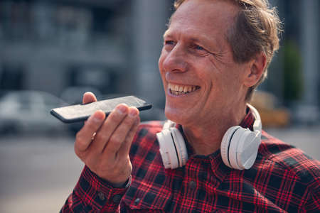 Close up of cheerful gentleman with headphone on his neck using speakerphone while talking on mobile...
