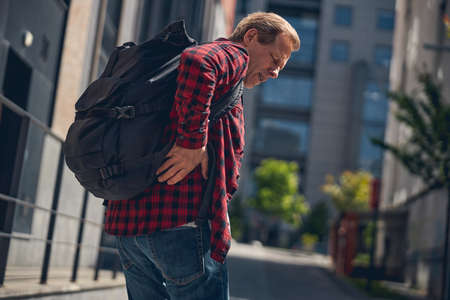 Middle-aged gentleman with backpack suffering from backache while standing on the streetの写真素材