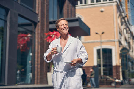 Handsome cheerful gentleman holding cup of coffee and smiling while strolling down the cityの写真素材