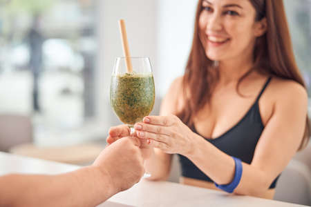 Smiling Caucasian lady with long dark hair receiving a vegetable drink in a beauty salonの写真素材
