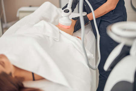 Cosmetologist doing cavitation procedure on a female client at a beauty center while woman lying