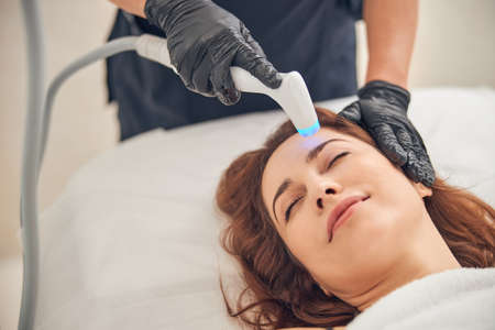 Attractive woman relaxing with closed eyes while getting procedure at beauty salon with help of cavitation deviceの写真素材