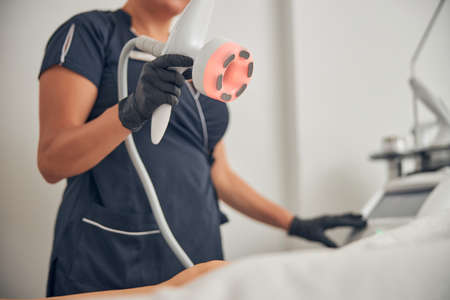Experienced cosmetologist in gloves preparing to use electronic equipment for improving skin of her patientの写真素材
