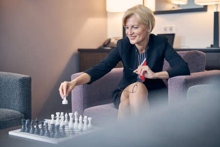 Beautiful blonde lady looking at chessboard and smiling while making opening move with white chess pawnの写真素材