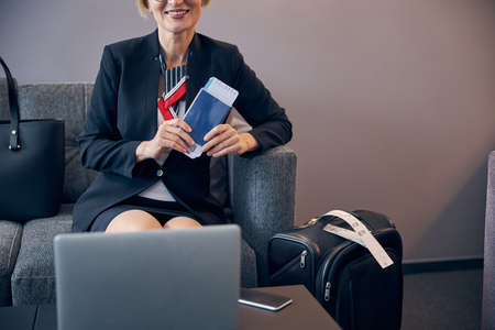 Elegant woman with airplane ticket in hand sitting on couch by the table with laptop and waiting for the flightの写真素材