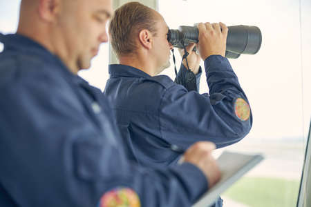 Waist up portrait of male looking through the binoculars while holding it in hands