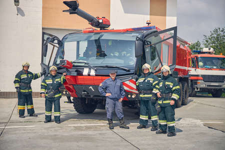 Full length portrait of happy firefighter team with equipment against trucks at fire station