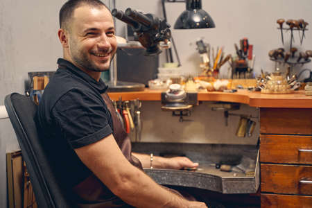 Waist-up portrait of a cheerful young male jeweler sitting in a chair at the workbench