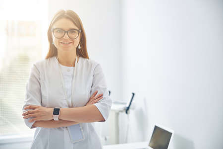 Smiling woman in white coat standing at the window with both hands on belly in medical clinic