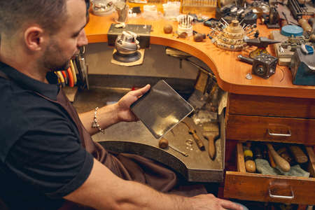 Side view of a dark-haired Caucasian male worker with a gadget sitting at the workbench