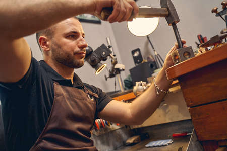 Concentrated young dark-haired Caucasian professional male jeweler shaping a metal workpiece in a...