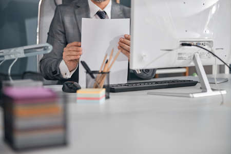 Close up of gentleman in stylish gray suit holding papers while sitting at the table with desktop PC...