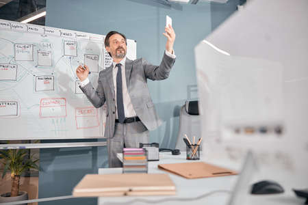 Handsome smiling man talking with colleague through video call while standing near whiteboard in...