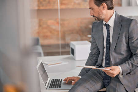 Serious gentleman in stylish suit holding documents and working on laptop while sitting on office...