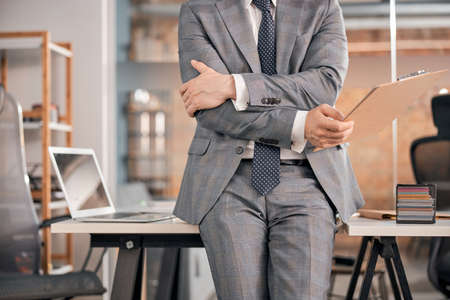 Close up of male worker in stylish gray suit holding clipboard and crossing arms