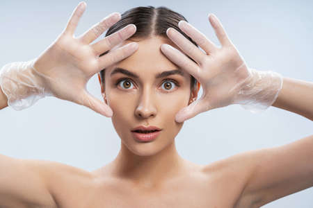 Close up of attractive amazed lady with perfect skin framing face with hands while standing against light blue backgroundの写真素材