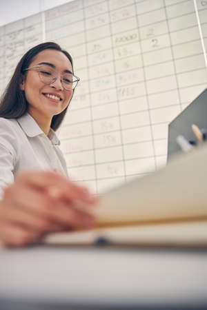 Charming brunette female person keeping smile on her face while sitting at her workplace
