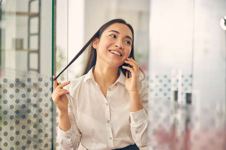 Attractive brunette female person keeping smile on her face while playing with hair during talkの写真素材