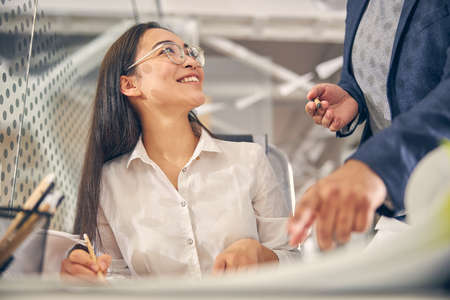 Kind brunette female person keeping smile on her face while making notes during consultationの写真素材