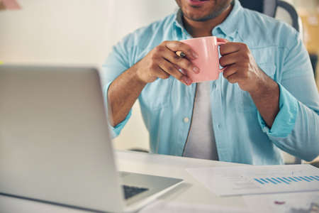 Concentrated young male person holding cup in both hands and looking at screen of his computer