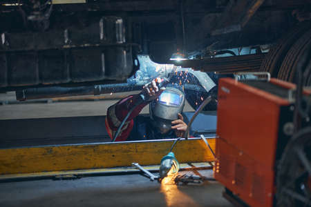 Busy welder standing under motor vehicle while working with mask during welding
