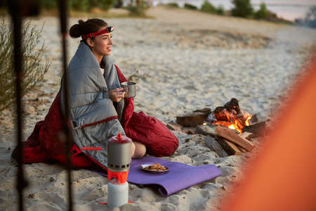 Beautiful young woman wrapped in sleeping bag holding cup of tea and smiling while sitting on sand...