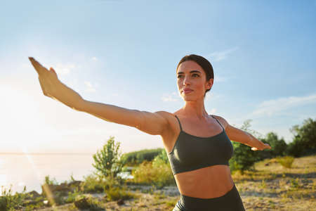 Charming lady in black crop top spreading hands to the sides while doing yoga exercise under...