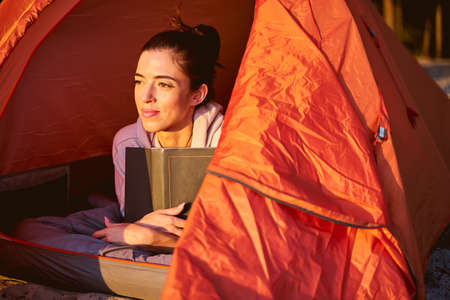 Charming female traveler with book looking away and smiling while lying on stomach inside touristic...