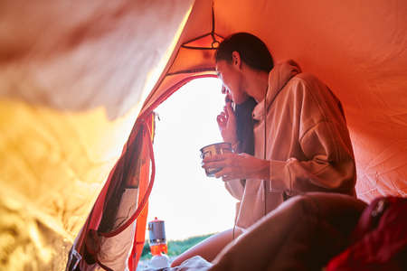 Charming female traveler holding mug of hot drink and eating cookie while sitting in touristic tentの写真素材