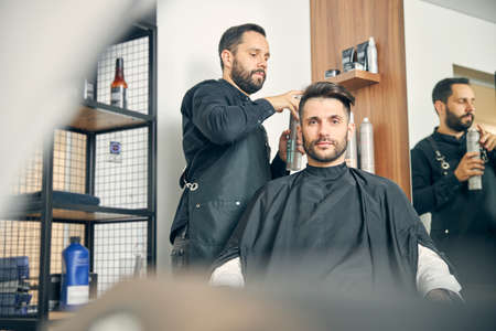Cheerful bearded man sitting in cozy armchair and looking forward while listening to his barberの写真素材