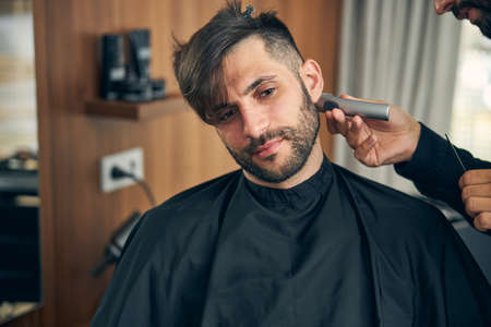 Handsome young male person sitting covered in cape while doing stylish haircutの写真素材