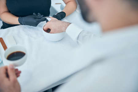 Kind brunette man preparing nails for manicure and enjoying his coffee in salonの写真素材