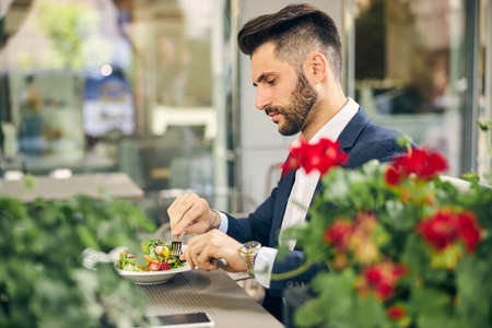 Attentive bearded man sitting in semi position and looking at colorful vegetables in plateの写真素材