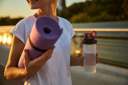 Close up of cheerful lady holding exercise mat and refreshing drink while standing on the streetの写真素材