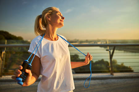 Charming sporty lady in white shirt holding jump rope and smiling while spending time outdoors