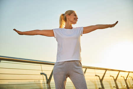 Beautiful lady in sportswear spreading hands to sides and smiling while doing exercise outdoors