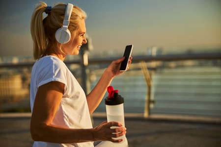Beautiful lady holding bottle of water and smiling while listening to music through wireless...