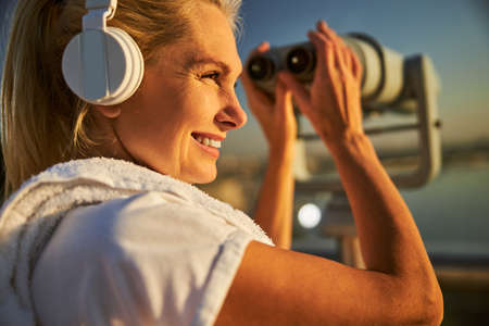 Charming lady listening to music and smiling while standing by stationery binoculars on observation...