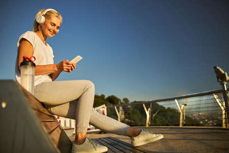 Charming lady holding cellphone and smiling while listening to music through wireless headphones and...