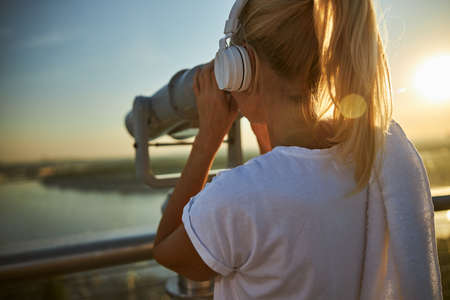 Close up of lady listening to music and looking at cityscape through stationery binoculars on observation deckの写真素材