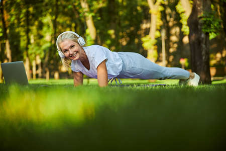 Charming lady in wireless headphones doing exercise and smiling while watching online training on...