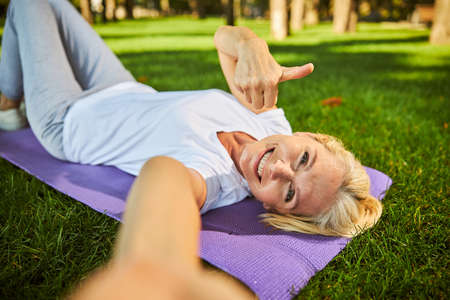 Beautiful lady showing approval gesture and smiling while resting on yoga mat in parkの写真素材