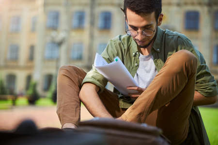 Focused short-haired bearded undergraduate in eyeglasses with papers in his hand looking down at somethingの写真素材