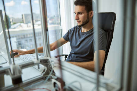 Serious focused dark-haired Caucasian young man in a t-shirt sitting in front of his computer